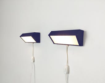 Puede incluir: Dos lámparas de pared de color azul oscuro con carcasas rectangulares y paneles de luz inclinados. Cada lámpara cuenta con un cable de alimentación blanco con un interruptor basculante blanco integrado que cuelga verticalmente.
