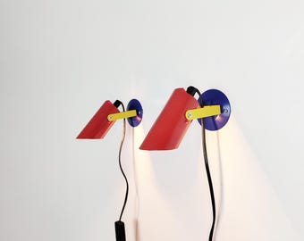 Puede incluir: Dos lámparas de pared con un diseño retro. Cada lámpara presenta una pantalla roja en forma de cono, un brazo amarillo y una base circular azul. Los cables negros se extienden desde las lámparas. Las lámparas están montadas en una pared blanca.