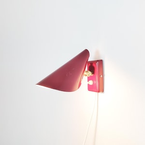 May include: A wall-mounted lamp featuring a conical, deep red metal shade attached to a matching rectangular base. The fixture includes a brass-coloured adjustment knob and a small white toggle switch, with a white power cord extending downwards.