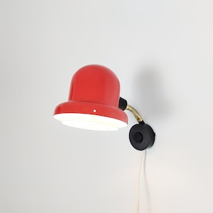 May include: A red, retro-style wall lamp. It has a rounded shade, a black and gold flexible arm, and a black base. The lamp is plugged into a wall and is turned on, emitting a bright white light.