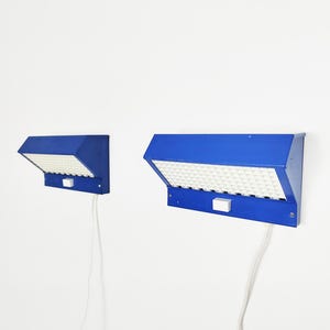 May include: Two blue wall lights with a geometric design. Each light has a rectangular shape with a white, textured surface and a small white switch. The lights are connected to a white cable.