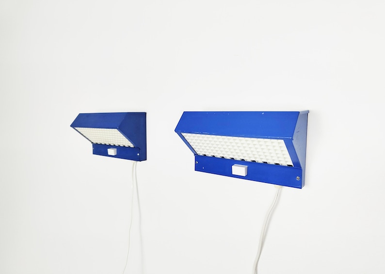 May include: Two blue wall lights with a geometric design. Each light has a rectangular shape with a white, textured surface and a small white switch. The lights are connected to a white cable.