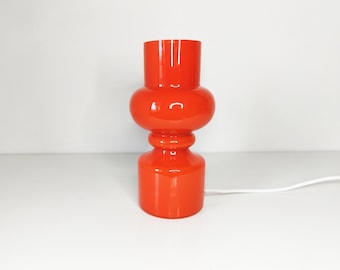 Könnte beinhalten: Eine leuchtend orangefarbene Tischleuchte aus Glas mit zylindrischem Fuß, einem abgerundeten Mittelteil und einer geraden oberen Öffnung. Die Lampe hat eine glatte, glänzende Oberfläche und ein weißes Stromkabel am Fuß.