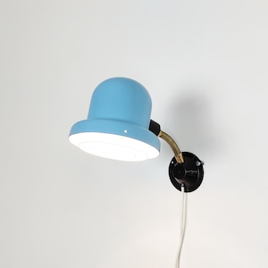 Può includere: Una lampada da parete azzurra con base nera e braccio flessibile color ottone. La lampada ha un paralume arrotondato e un interno bianco. Un cavo bianco si estende dalla base nera.