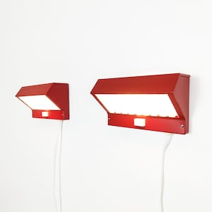 May include: Two red metal wall lamps with angled shades and white rectangular light diffusers. Each lamp features a small white toggle switch on the front panel and a white power cord extending downward from the base against a plain white wall.