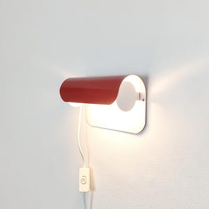 Könnte beinhalten: Eine Wandleuchte mit einem geschwungenen, matten roten Metallschirm über einer weißen, rechteckigen Rückplatte. Das Licht strahlt warm unter dem Schirm hervor, und ein weißes Stromkabel mit einem rechteckigen Schalter hängt an der Leuchte.