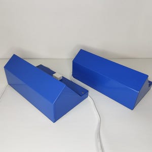 May include: Two blue geometric lamps with white cords. The lamps have a rectangular base and a slanted top. The lamps are made of a glossy material and are placed on a white surface.
