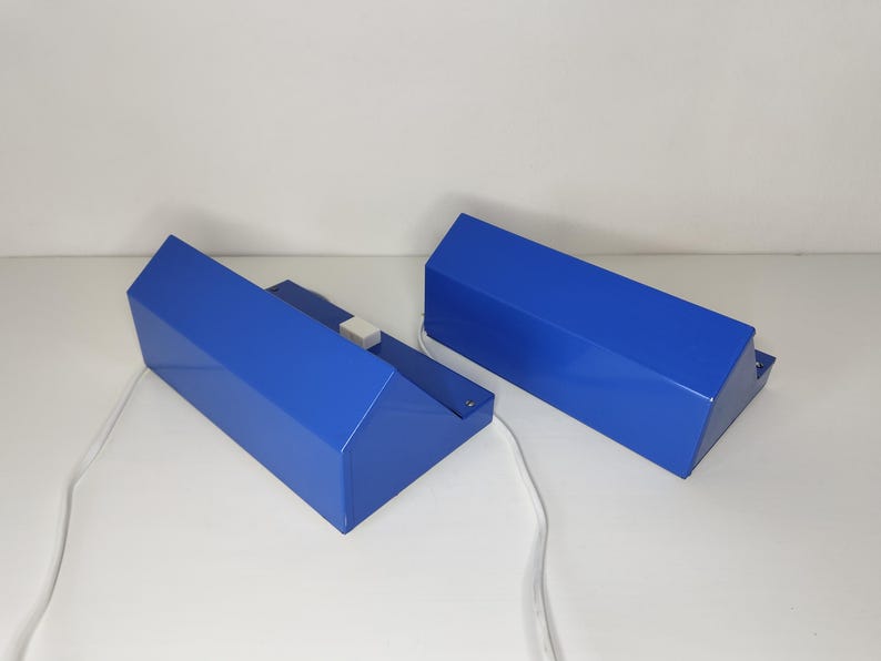 May include: Two blue geometric lamps with white cords. The lamps have a rectangular base and a slanted top. The lamps are made of a glossy material and are placed on a white surface.