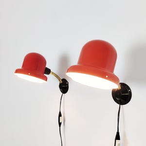 Könnte beinhalten: Zwei Wandleuchten mit glänzenden orangefarbenen kuppelförmigen Schirmen, schwarzen runden Wandplatten und schwarzen Stromkabeln mit Zwischenschaltern. Die kleinere Leuchte hat einen gebogenen Messingarm, die größere ist fest montiert.