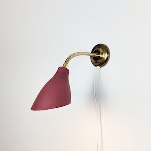 Può includere: Una lampada da parete con paralume bordeaux e braccio flessibile color ottone. La lampada ha una base circolare in ottone e un cavo di alimentazione bianco. Il paralume è di colore rosso scuro e la lampada è montata su una parete bianca.