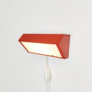 Può includere: Una lampada da parete rosso brillante con un alloggiamento in metallo rettangolare e un pannello diffusore bianco con motivo a griglia. L'apparecchio include un cavo di alimentazione bianco con un interruttore a bilanciere integrato.