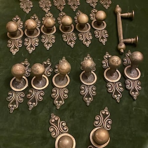 Op de afbeelding: Een assortiment antieke messing hardware, waaronder deurknoppen en -trekkers, gerangschikt op een groene fluwelen ondergrond. De hardware heeft sierlijke, krullende ontwerpen en een warme, verouderde patina, wat een vintage esthetiek suggereert.