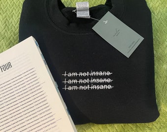 Pode incluir: Uma camisola preta de gola redonda com texto bordado a branco que diz "I am not insane" repetido três vezes, com uma linha horizontal a atravessar cada frase. Uma etiqueta cinzenta retangular está presa à gola com o texto "malis apparel".