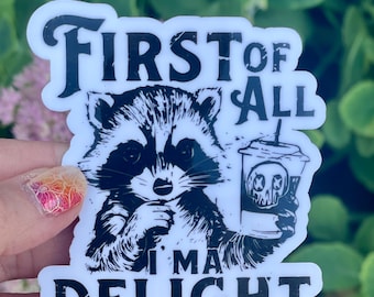 Puede incluir: Una pegatina blanca con una ilustración en blanco y negro de un mapache sosteniendo una taza de café. El texto en la pegatina dice "First of all I'm a Delight".