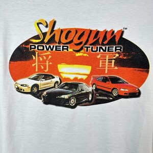 May include: White t-shirt with a red, orange, and yellow oval graphic. The graphic features the text "Shogun Power Tuner" and three cars: a white Mitsubishi Eclipse, a black Honda Civic, and a red Honda Civic. The graphic also includes Japanese characters.