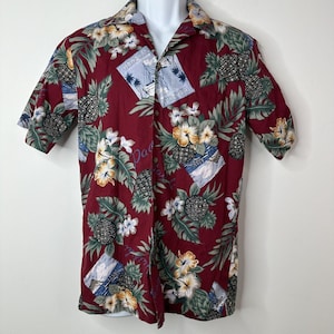 Peut inclure: Une chemise à manches courtes bordeaux avec un imprimé tropical. Le motif présente des ananas, des fleurs et des voiliers sur un fond de feuilles vertes. Le mot "Paradise" est imprimé sur la chemise.