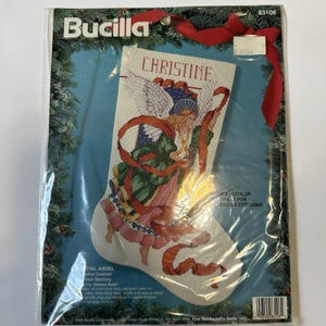 May include: A Bucilla Christmas stocking kit featuring a cross-stitch design of an angel with wings, holding a ribbon. The stocking is white with a colorful angel design, and the name "CHRISTINE" is at the top.