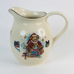 Puede incluir: Una jarra de cerámica de color crema con un borde verde y un diseño navideño con un oso de peluche que lleva un gorro de Papá Noel y sostiene un regalo. El oso está rodeado de regalos y estrellas.