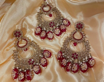 May include: A set of red and gold Indian bridal jewelry, including a maang tikka and earrings. The earrings feature a large, ornate design with red stones and pearls. The maang tikka is a similar design, but smaller. The jewelry is set against a gold fabric background.