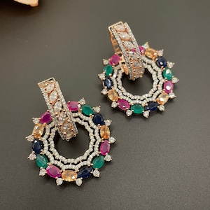 May include: A pair of gold-toned earrings with a circular design. The earrings feature a variety of colorful gemstones, including pink, green, blue, and orange.