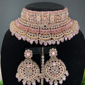 May include: A pink and gold Indian bridal necklace set with a choker, earrings, and a maang tikka. The set is adorned with clear and pink stones, pearls, and beads.
