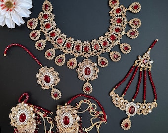 May include: A traditional Indian bridal jewelry set with a gold necklace, earrings, a maang tikka, and a hathphool. The set features red and clear stones.