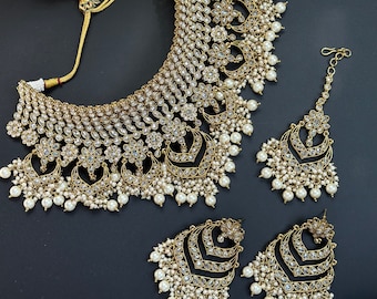 Puede incluir: Un conjunto de joyas en tono dorado con collar, pendientes y tocado. El collar y los pendientes tienen un diseño en capas con piedras transparentes y perlas de imitación colgantes. El tocado tiene un diseño floral central y una gota de perla.