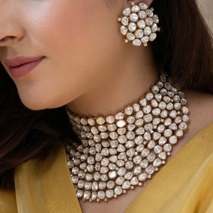 May include: A gold choker necklace featuring a large, elaborate design crafted from small, clear, faceted stones. The necklace is worn by a person with dark hair and a fair complexion.