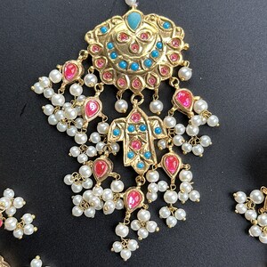 May include: An Indian bridal headpiece in gold tones, featuring a large, ornate centrepiece and dangling pearl and pink and blue gemstone accents.