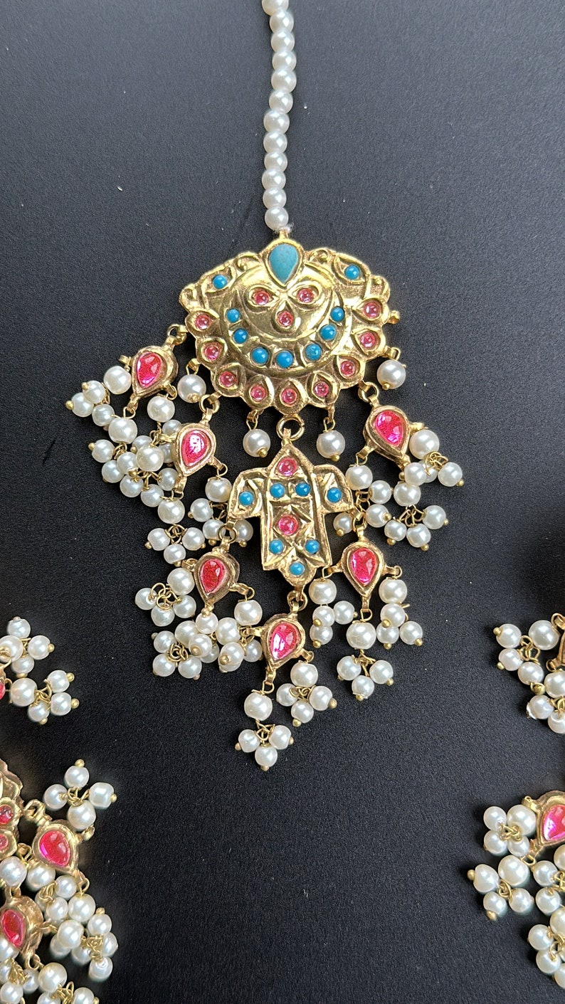 May include: An Indian bridal headpiece in gold tones, featuring a large, ornate centrepiece and dangling pearl and pink and blue gemstone accents.