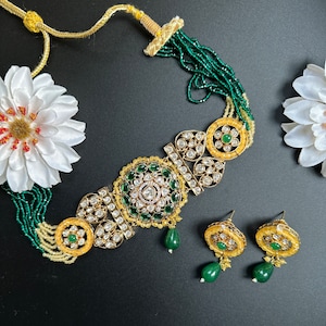 Peut inclure: Un collier en or avec des pierres vertes et blanches, et des boucles d'oreilles assorties. Le collier a une chaîne perlée et un grand pendentif de forme circulaire. Les boucles d'oreilles sont petites et ont un design similaire au pendentif.