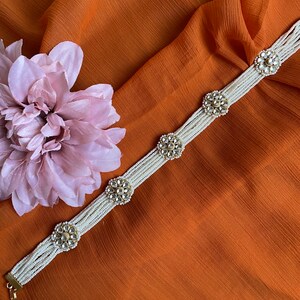 May include: A white pearl and gold choker necklace with four decorative round floral accents. The necklace is on a brown fabric background.