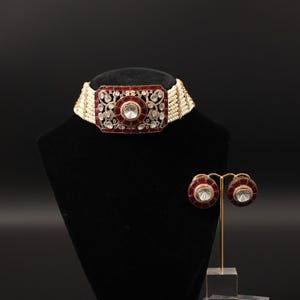 May include: A red and white beaded choker necklace with matching earrings. The necklace features a large rectangular centerpiece with a central clear stone and surrounding smaller stones. The earrings are round with a similar design.