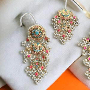 May include: A pair of gold earrings with pink and blue gemstones, pearl accents, and a decorative design. The earrings are on a white fabric background.