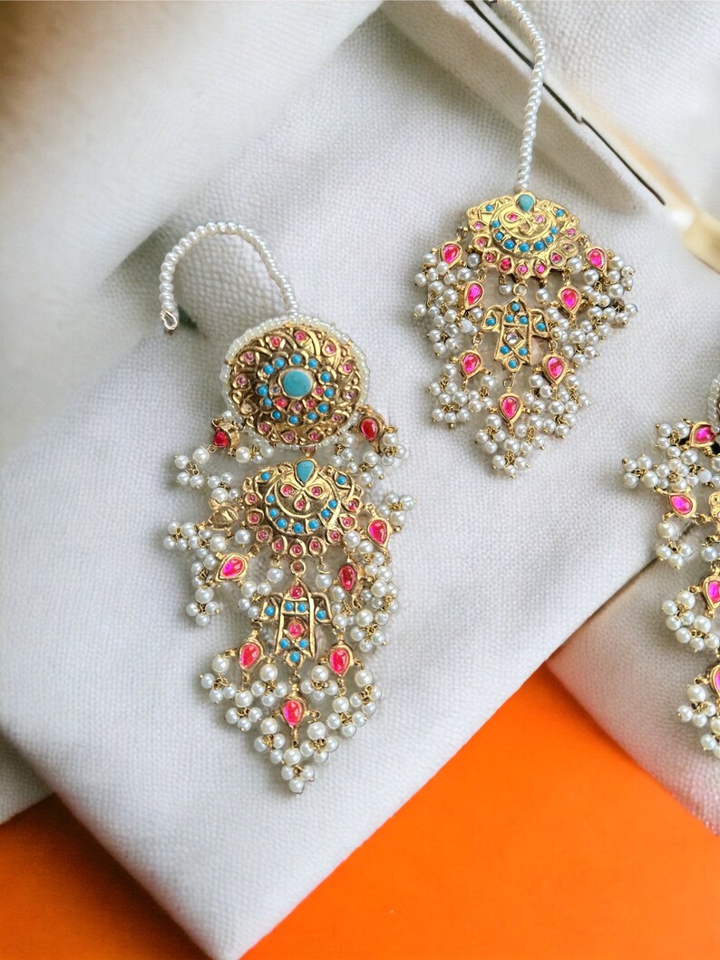 May include: A pair of gold earrings with pink and blue gemstones, pearl accents, and a decorative design. The earrings are on a white fabric background.