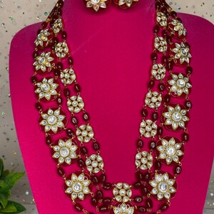 May include: A four-strand layered necklace with red beads and gold-toned round pendants featuring white stones. The necklace is displayed on a pink mannequin bust. Matching earrings are also shown.