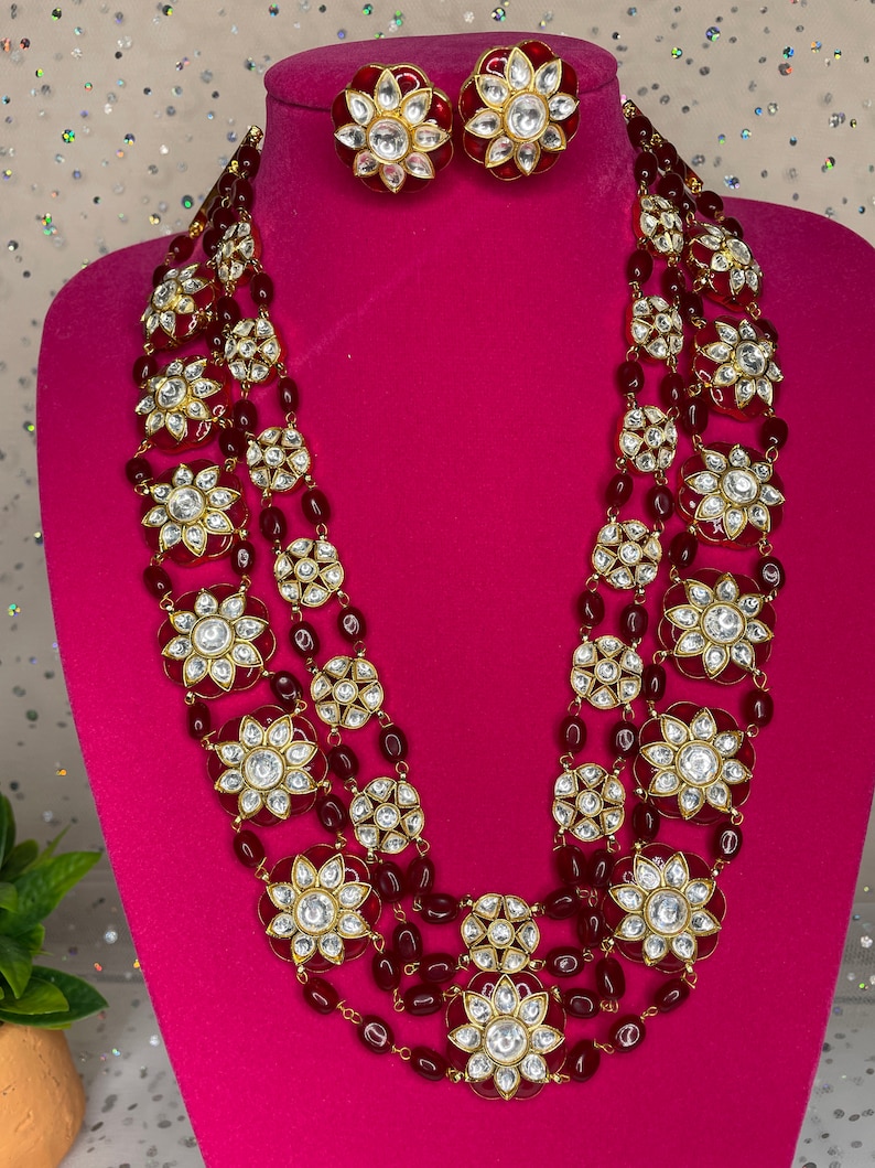 May include: A four-strand layered necklace with red beads and gold-toned round pendants featuring white stones. The necklace is displayed on a pink mannequin bust. Matching earrings are also shown.