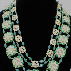 May include: A three-strand necklace with green and gold beads and white flower-shaped pendants. The necklace is made of metal and has a traditional Indian design.