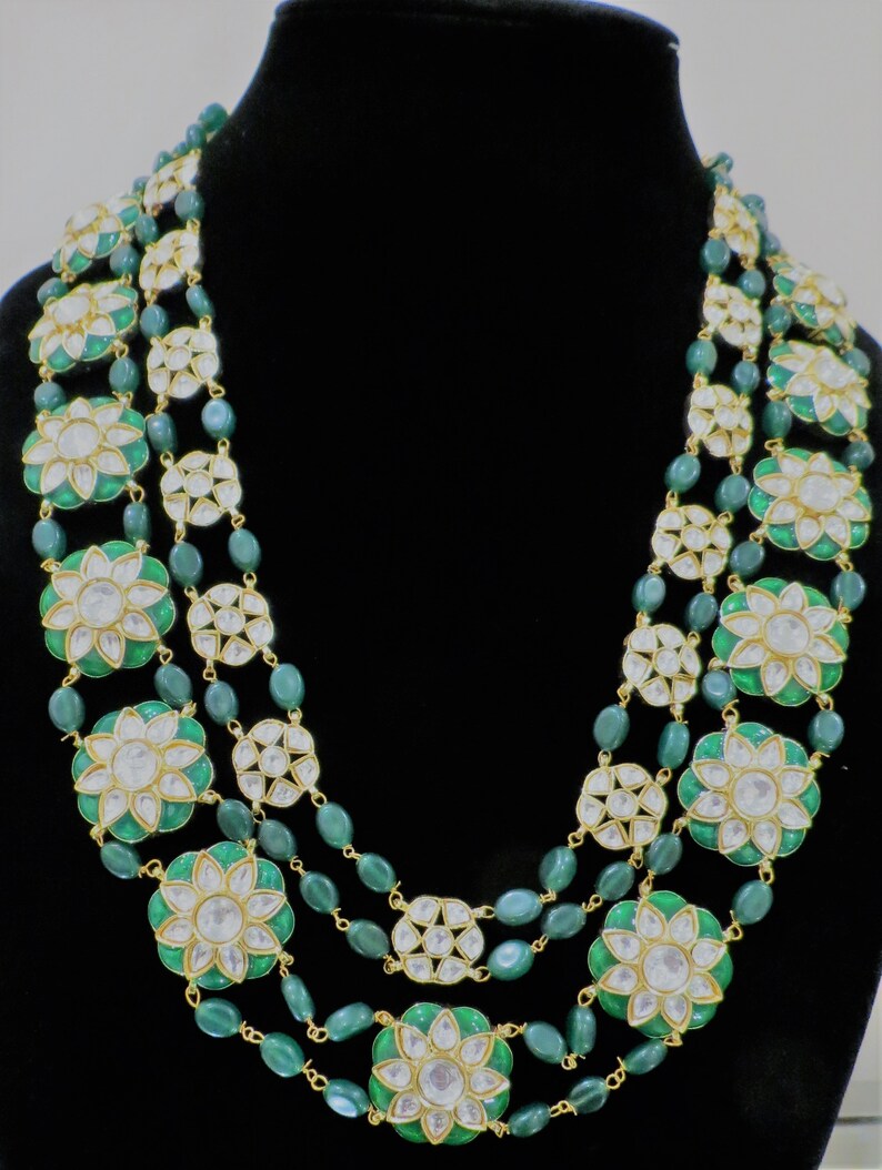 May include: A three-strand necklace with green and gold beads and white flower-shaped pendants. The necklace is made of metal and has a traditional Indian design.