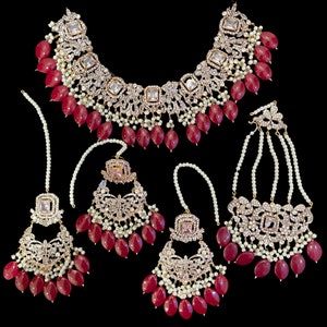 May include: A traditional Indian bridal jewelry set featuring a necklace, earrings, and a maang tikka. The set is made of gold-toned metal and features intricate designs with clear stones and red beads.