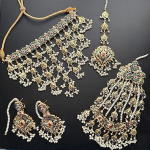 May include: A gold-toned jewelry set featuring a necklace, earrings, and headpieces. The set includes intricate designs with colorful accents, small pearls, and dangling elements. The jewelry is displayed on a dark surface, highlighting the details and craftsmanship.