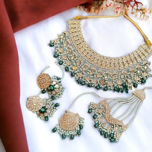 May include: A gold and green Indian bridal necklace set with pearl and emerald accents. The necklace features a large, ornate pendant with intricate details. The earrings are round and have a similar design to the pendant.