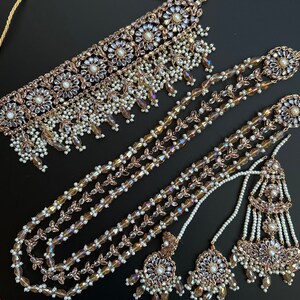 May include: A gold and white bridal necklace set with a choker and long layered necklace with matching earrings. The set is adorned with iridescent crystals and pearls.