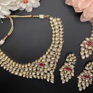 May include: A gold necklace and earrings set featuring a pink stone and clear rhinestones. The necklace is a choker style with a teardrop shape. The earrings are dangle style with a teardrop shape.