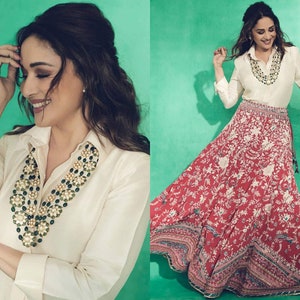 May include: A woman wearing a white button-down shirt with a long, layered green and gold necklace. She is also wearing a red and white floral print skirt.