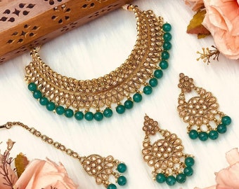 May include: A gold necklace and earrings set with green beads and intricate detailing. The necklace has multiple layers of gold with small stones and the earrings are teardrop shaped with a round center.