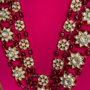 May include: A multi-strand necklace with red beads and gold-toned floral pendants with clear stones. The necklace is on a pink background.