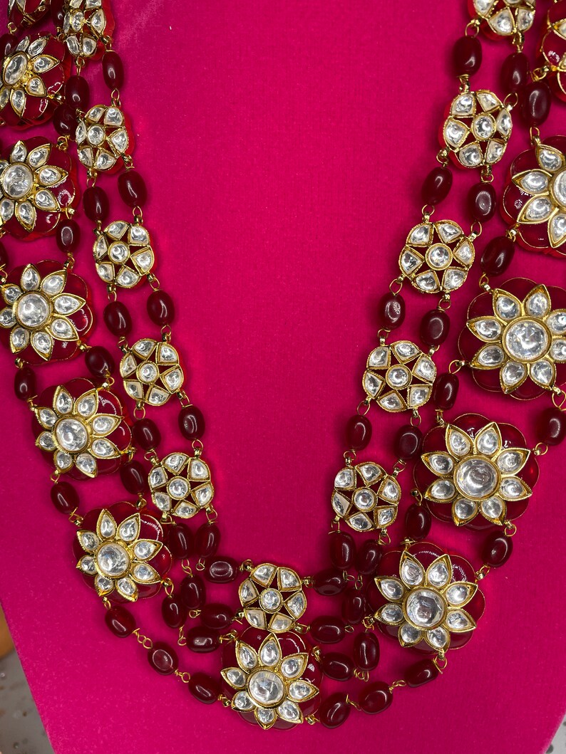 May include: A multi-strand necklace with red beads and gold-toned floral pendants with clear stones. The necklace is on a pink background.