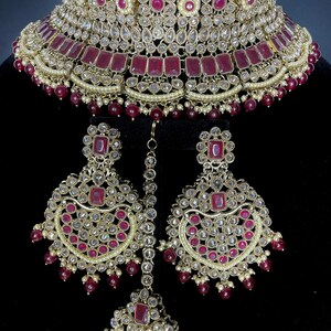 May include: A gold and maroon bridal necklace set with a choker style necklace, matching earrings, and a tikka. The set is adorned with clear and red stones.