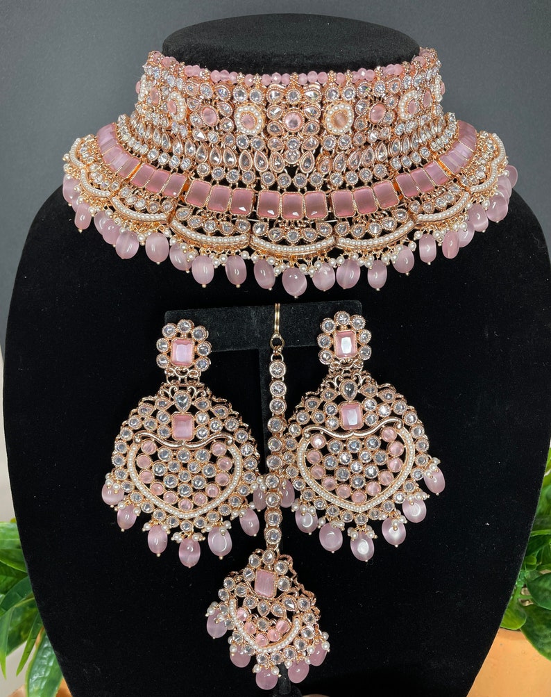 May include: A pink and gold Indian bridal necklace set with a choker, earrings, and a maang tikka. The set is adorned with clear and pink stones, pearls, and beads.